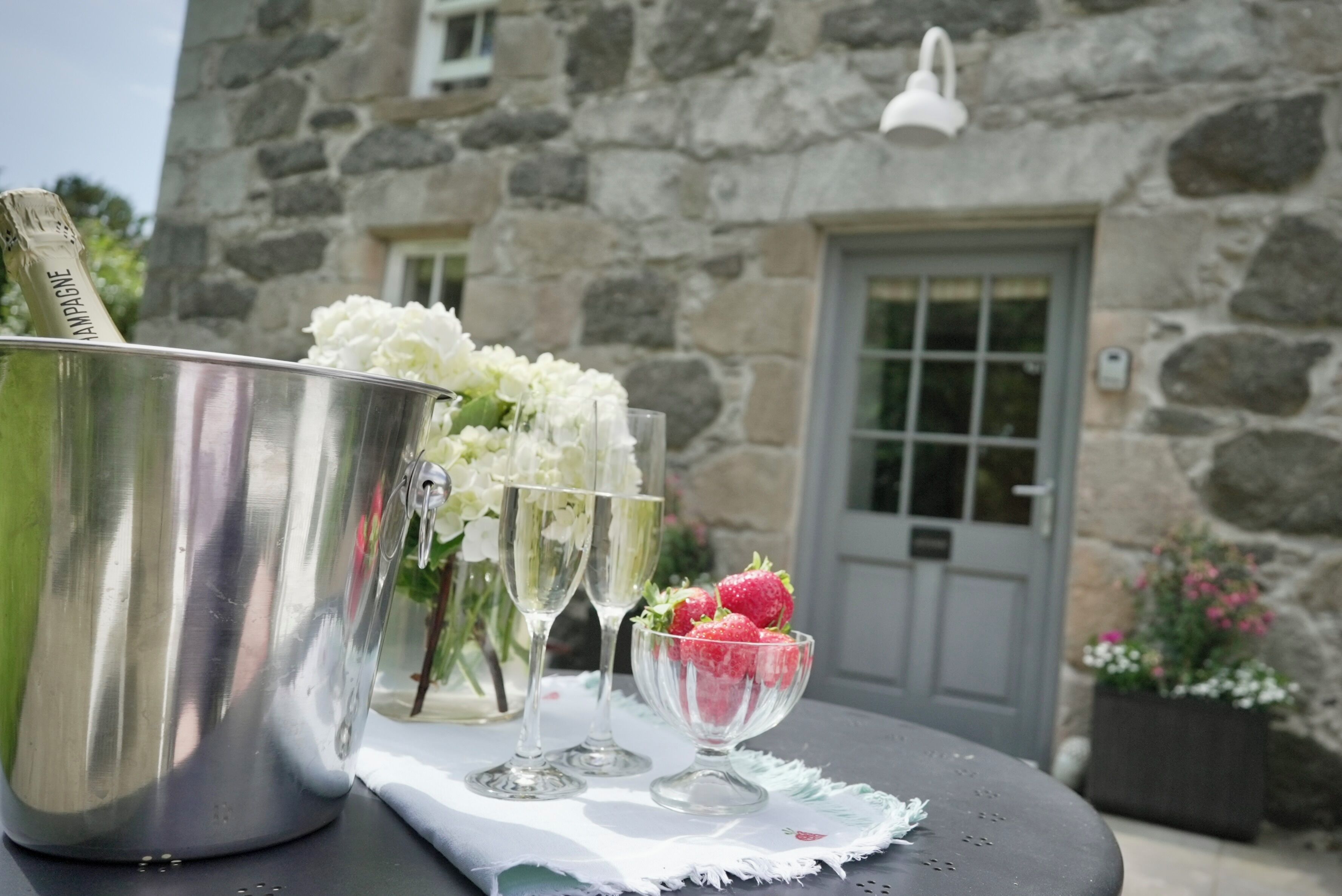 Un cubo de acero inoxidable para champán, dos copas de vino, un cuenco de fresas y flores blancas sobre una mesa frente a un edificio de piedra con puerta y ventana.