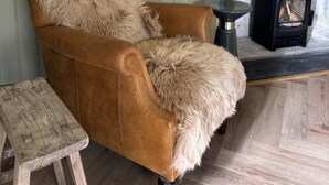 A brown leather armchair with a fur throw, a small wooden stool, and a black wood-burning stove against a wall with vertical paneling.