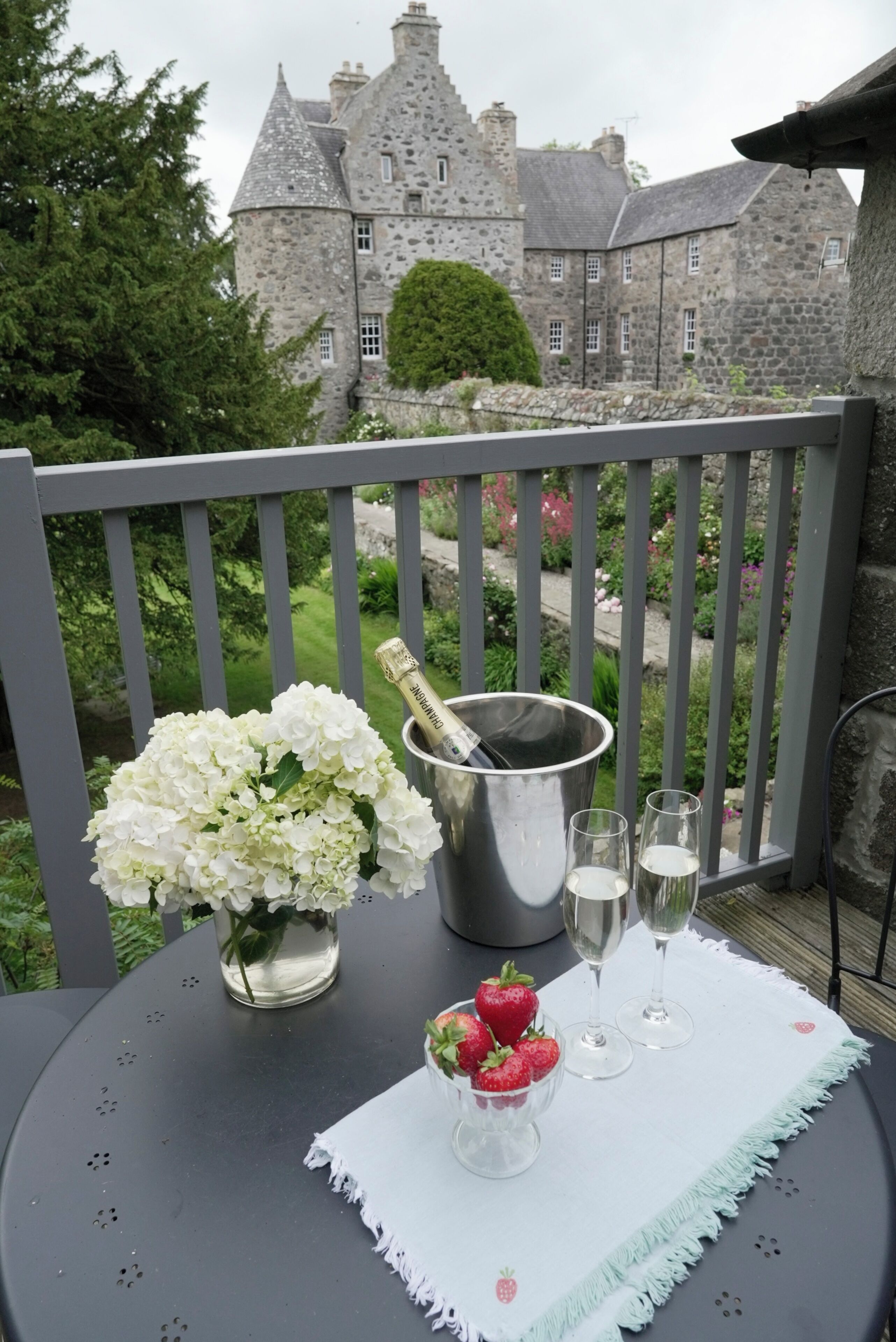 Un balcón con una mesa dispuesta para tomar algo, un ramo de flores y vistas a un castillo.