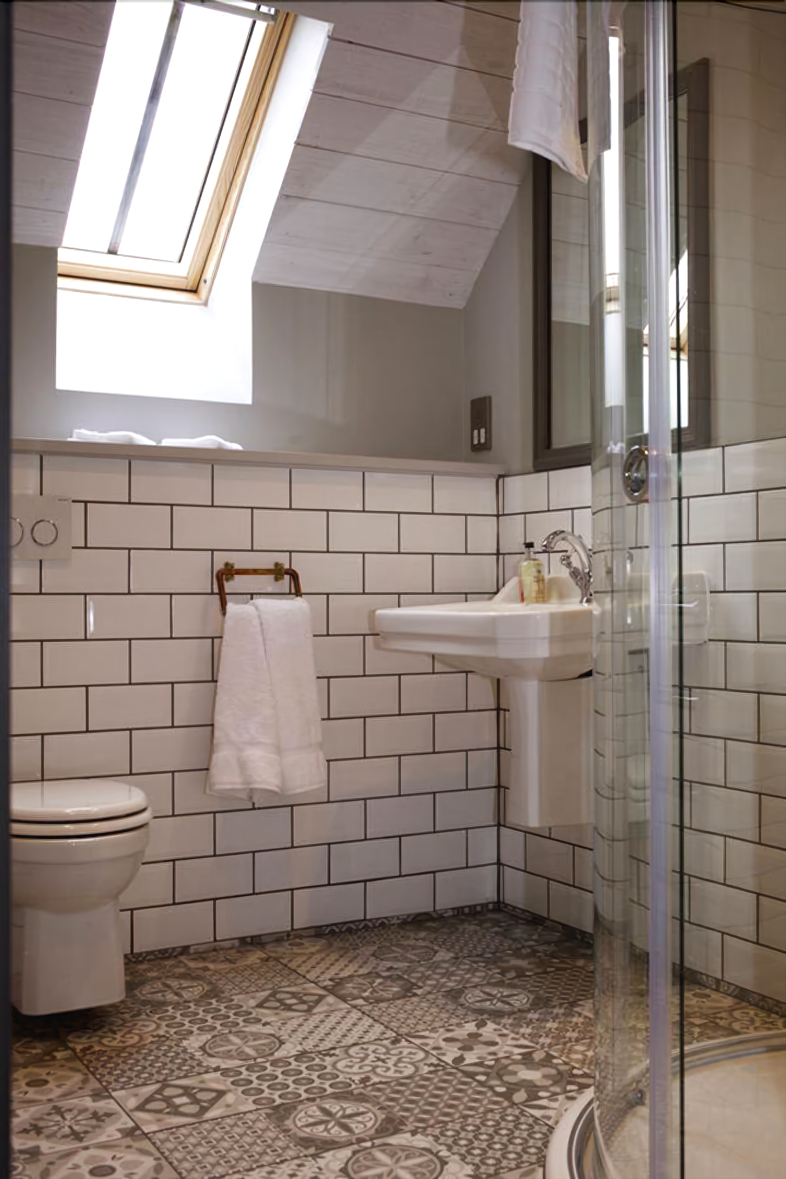 Un baño con tragaluz, paredes de azulejos blancos, un lavamanos, un inodoro y baldosas de suelo estampadas.