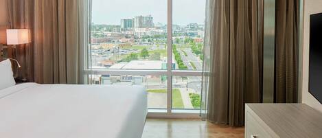 A hotel room with a large bed, a dresser, and a TV. There is a window with curtains and a view of a cityscape.