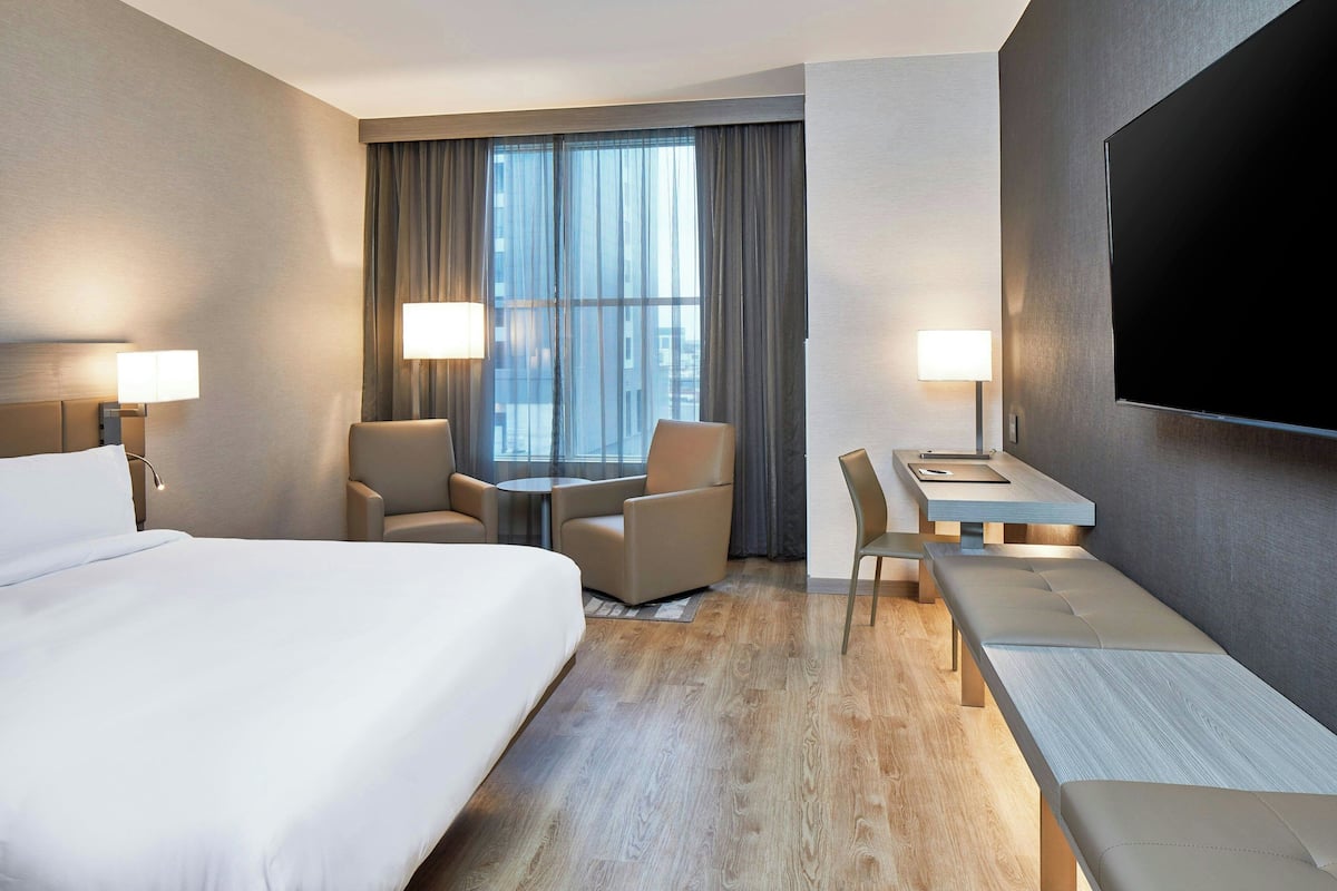 Ein Hotelzimmer mit einem großen Bett, zwei Sesseln, einem kleinen Tisch, einem Schreibtisch und einem an der Wand befestigten Fernseher.