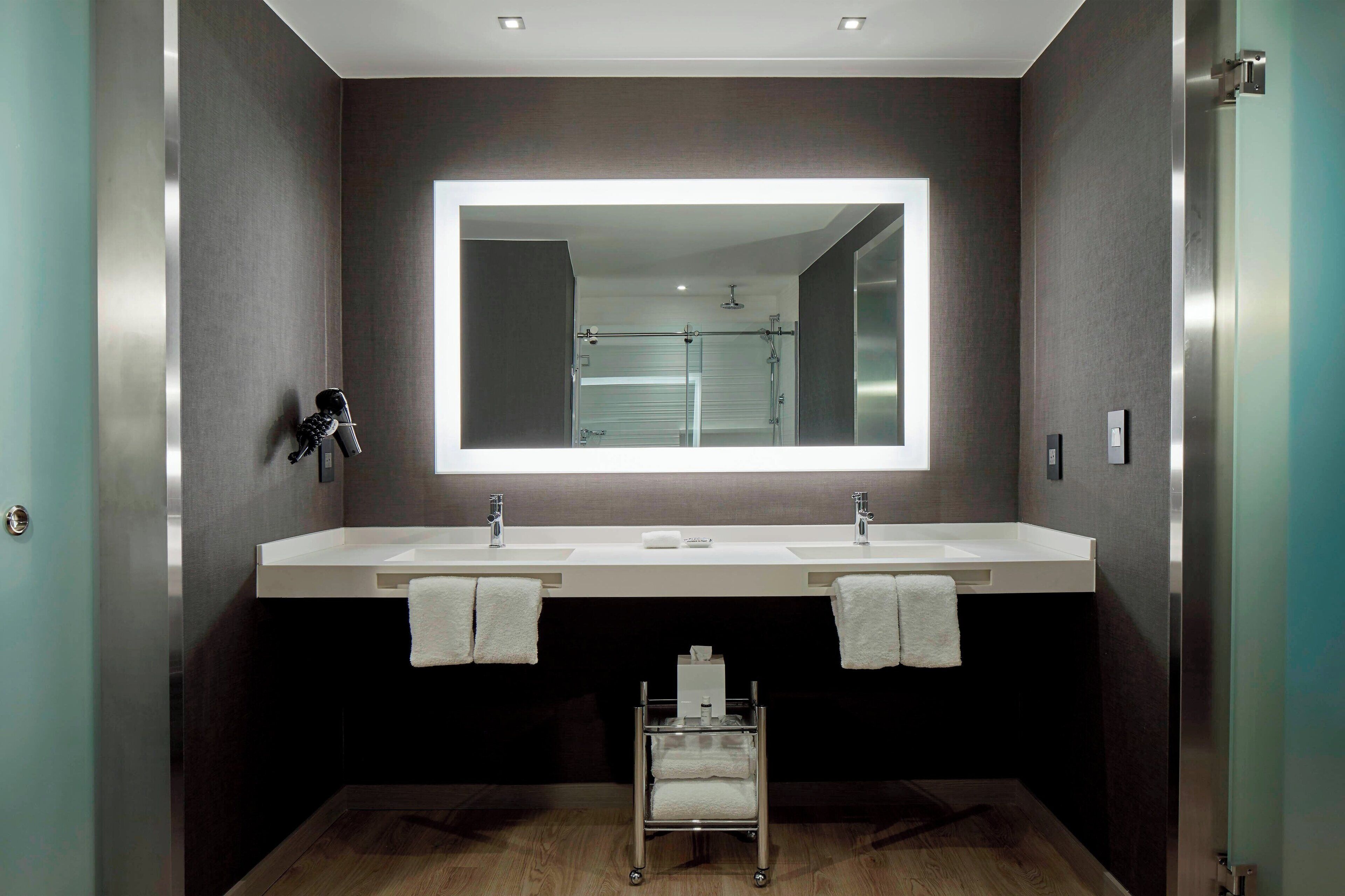 A modern bathroom with two sinks, a large mirror, and a small cart with towels.