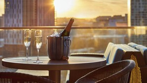 Una mesa con una botella de champán y dos copas en un balcón con vistas a un paisaje urbano al atardecer.