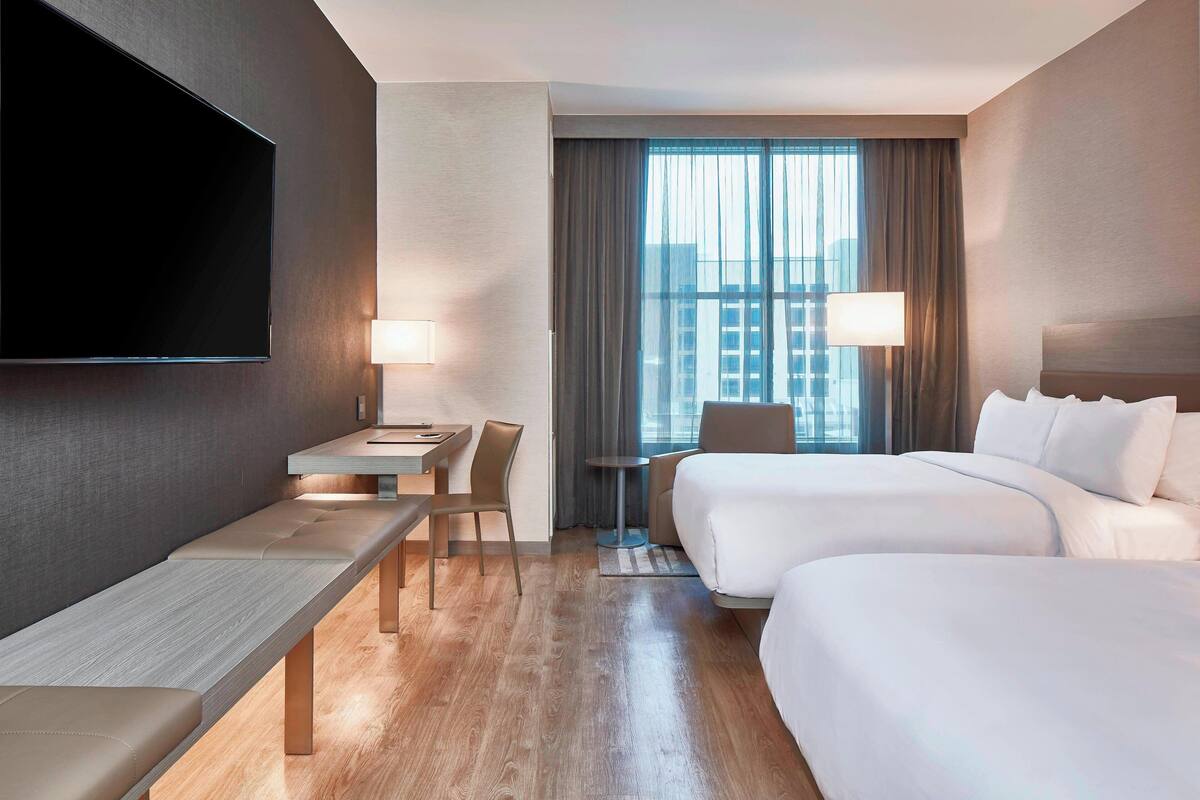 Ein Hotelzimmer mit Flachbildfernseher, Schreibtisch mit Stuhl, zwei Betten und einem Fenster mit Vorhängen.