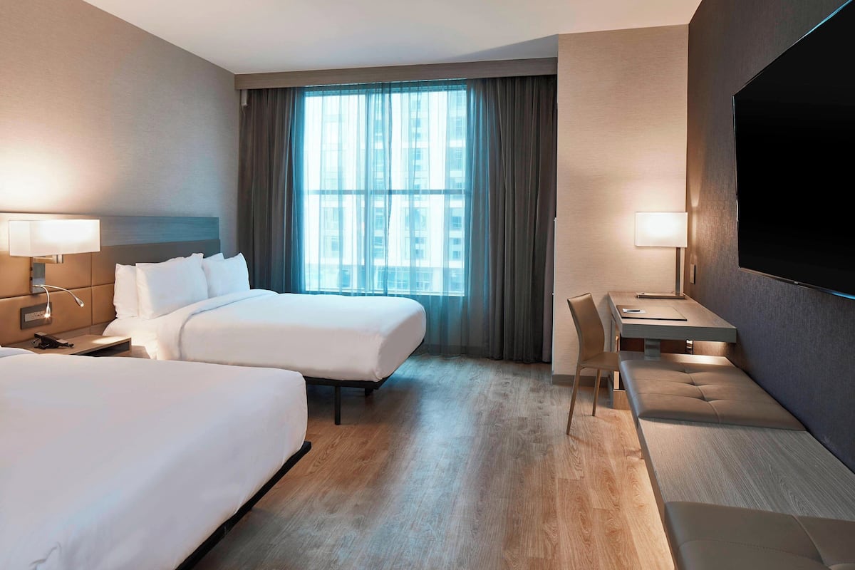 Ein Hotelzimmer mit zwei Betten, einem Schreibtisch, einem Stuhl, einem Fernseher und einem großen Fenster mit Vorhängen.