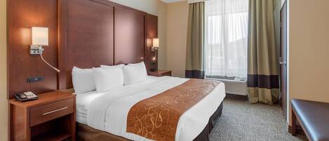 A hotel room with a large bed, wooden headboard, two bedside tables with lamps, a desk, and a window with curtains.
