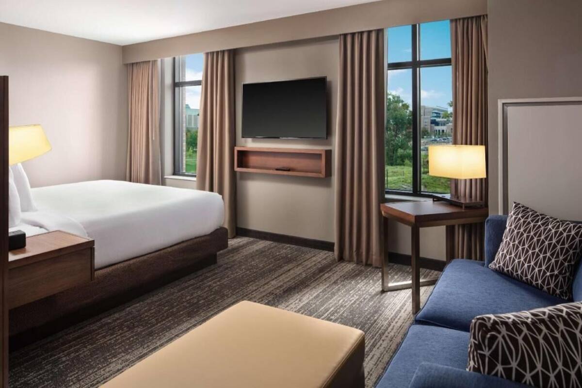 Ein Hotelzimmer mit Bett, Fernseher, Schreibtisch, Sofa und einem fensterbepflanzten Fenster.