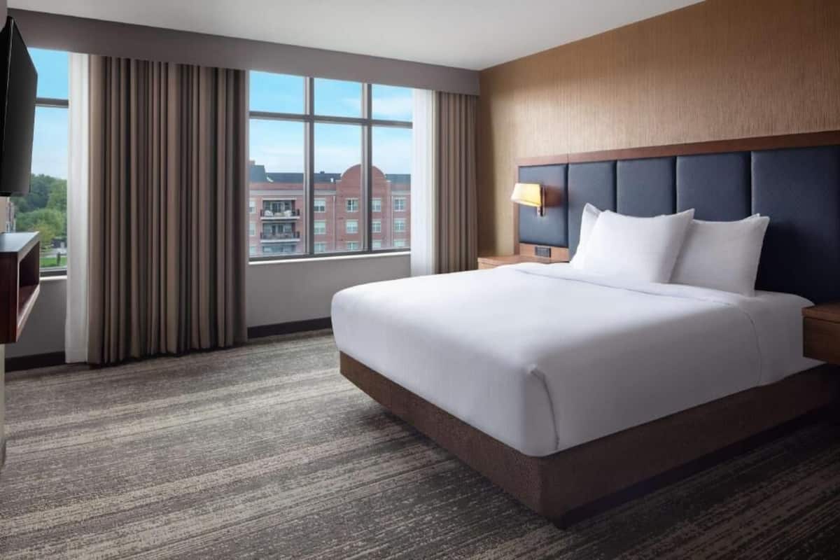 Ein Hotelzimmer mit einem großen Bett, einem Fernseher, einem Schreibtisch und Blick auf Gebäude.