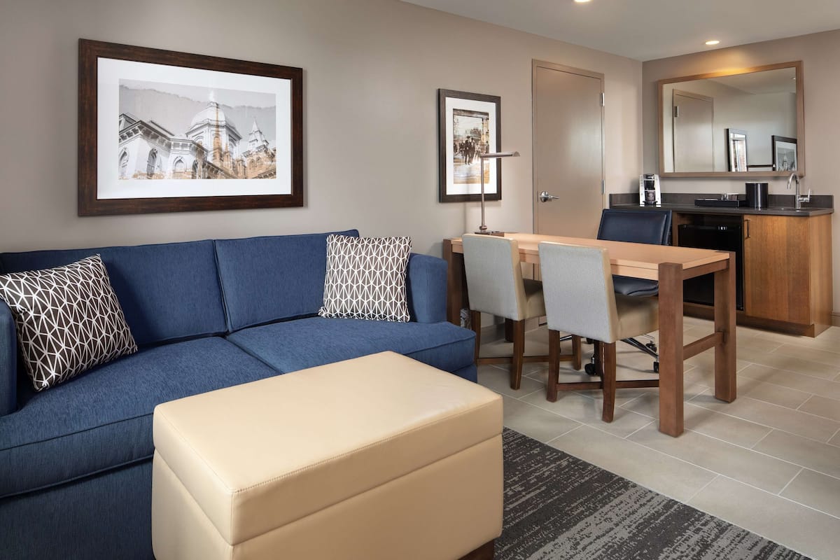 Ein modernes Wohnzimmer mit einem blauen Sofa, einem beigen Hocker und einem Essbereich mit einem Holztisch und Stühlen.
