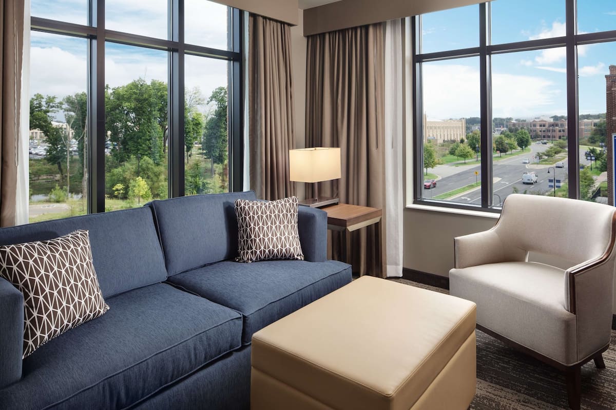Ein Hotelzimmer mit einem blauen Sofa, einem beige-farbenen Hocker und einem weißen Sessel. Ein Fenster bietet Blick auf eine Stadtansicht und Grünflächen.