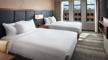 A hotel room with two beds, a TV, a desk, and a window with a view of buildings.