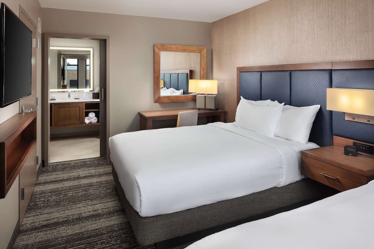 Ein Hotelzimmer mit einem großen Bett, einem Nachttisch mit Lampe, einem Spiegel, einem Fernseher und einem Badezimmer mit Waschbecken und Handtuchhalter.