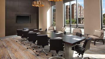 A modern conference room with a large table, multiple chairs, a flat-screen TV, and large windows offering a view of buildings outside.