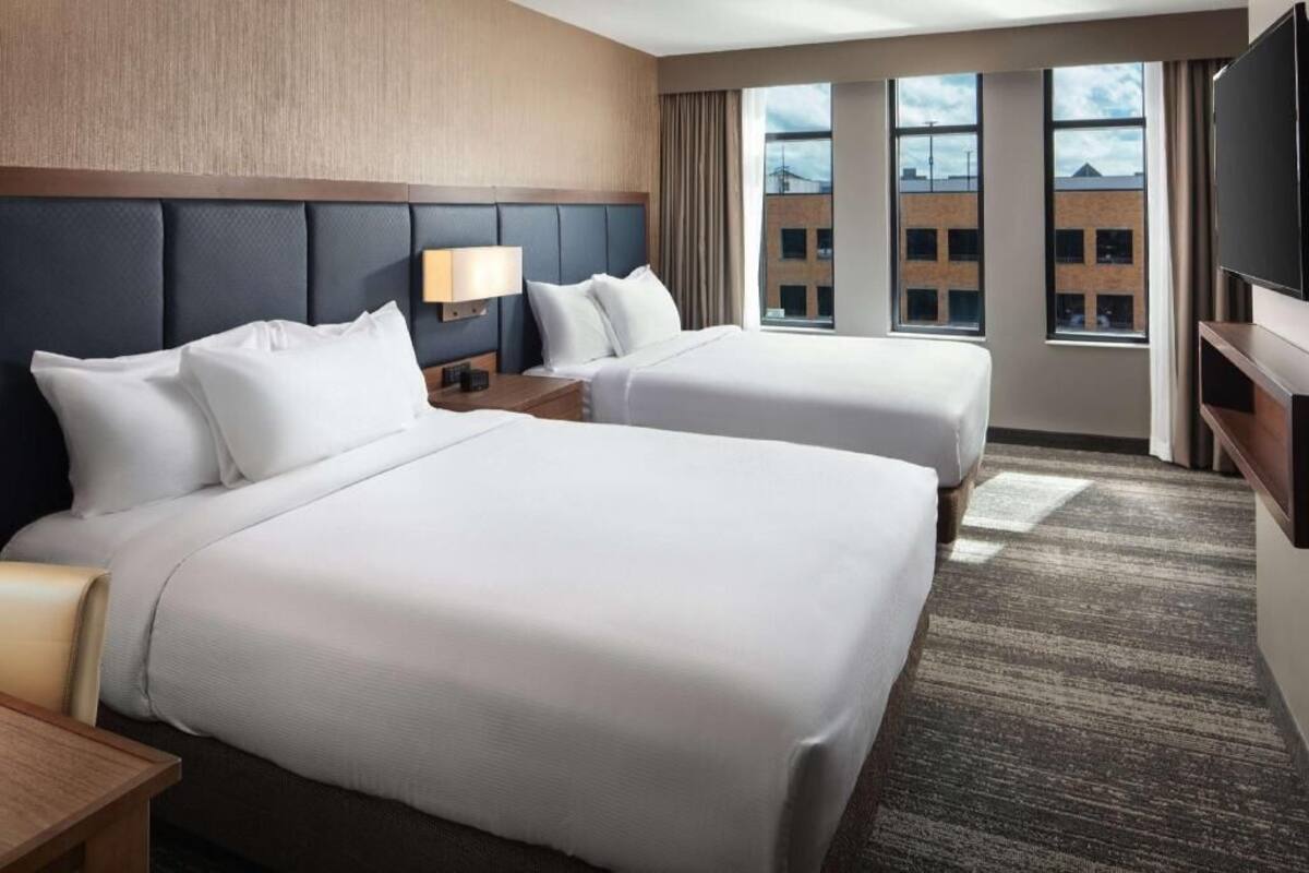 Ein Hotelzimmer mit zwei Betten, einem Fernseher, einer Lampe und Blick auf Gebäude.
