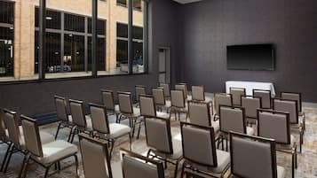 Un salón de conferencias con filas de sillas, un ventanal grande y un televisor de pantalla plana en la pared.