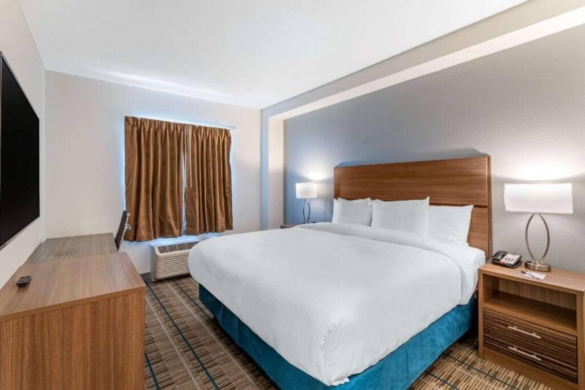 A hotel room with a bed, bedside table, lamp, air conditioning unit, and a TV.