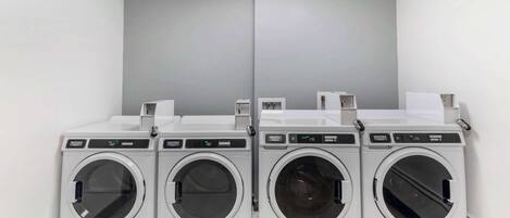 A row of four white front-loading washing machines in a laundry room with grey walls and a tiled floor.