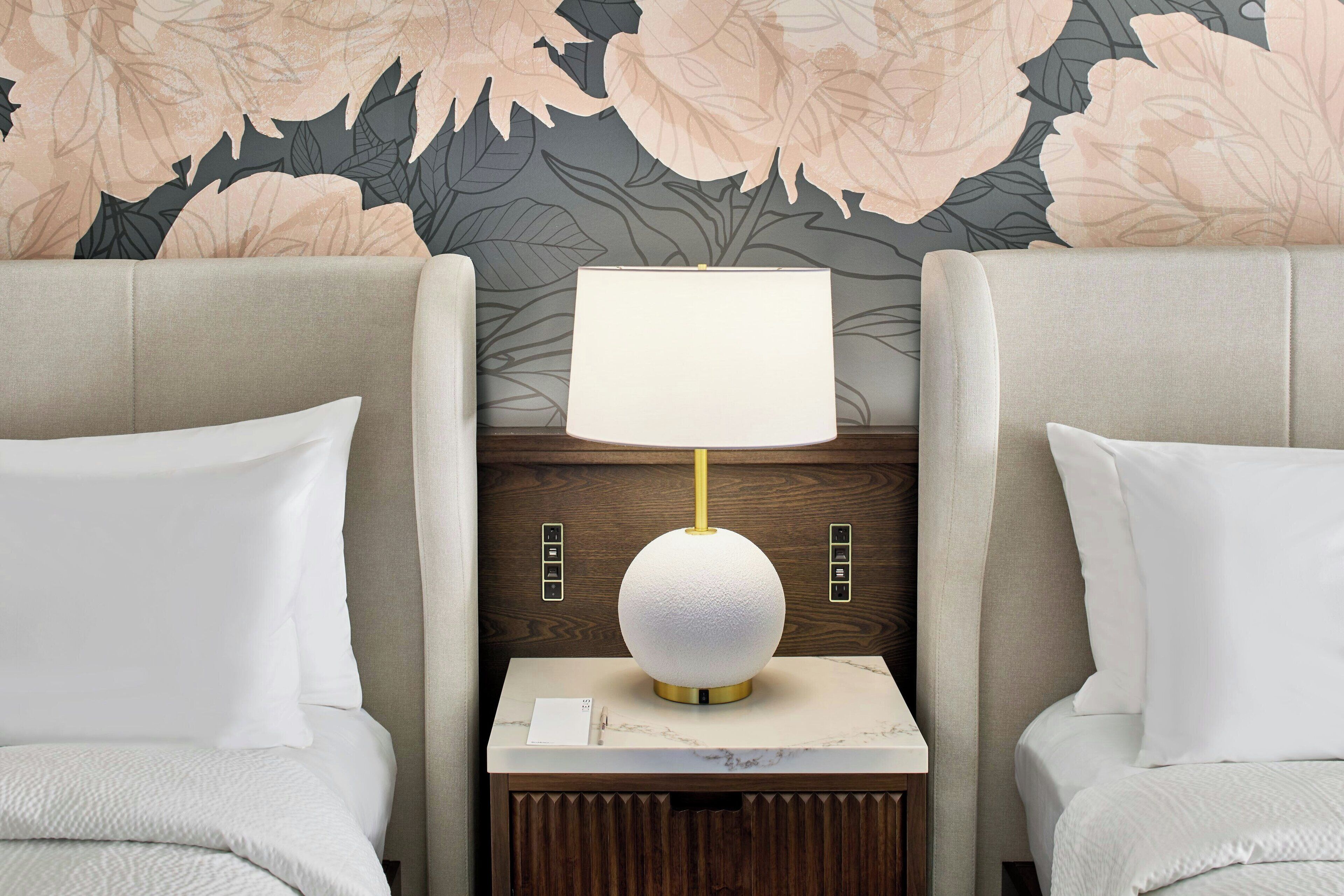 Camera d'albergo con due letti, un comodino con una lampada e carta da parati floreale.