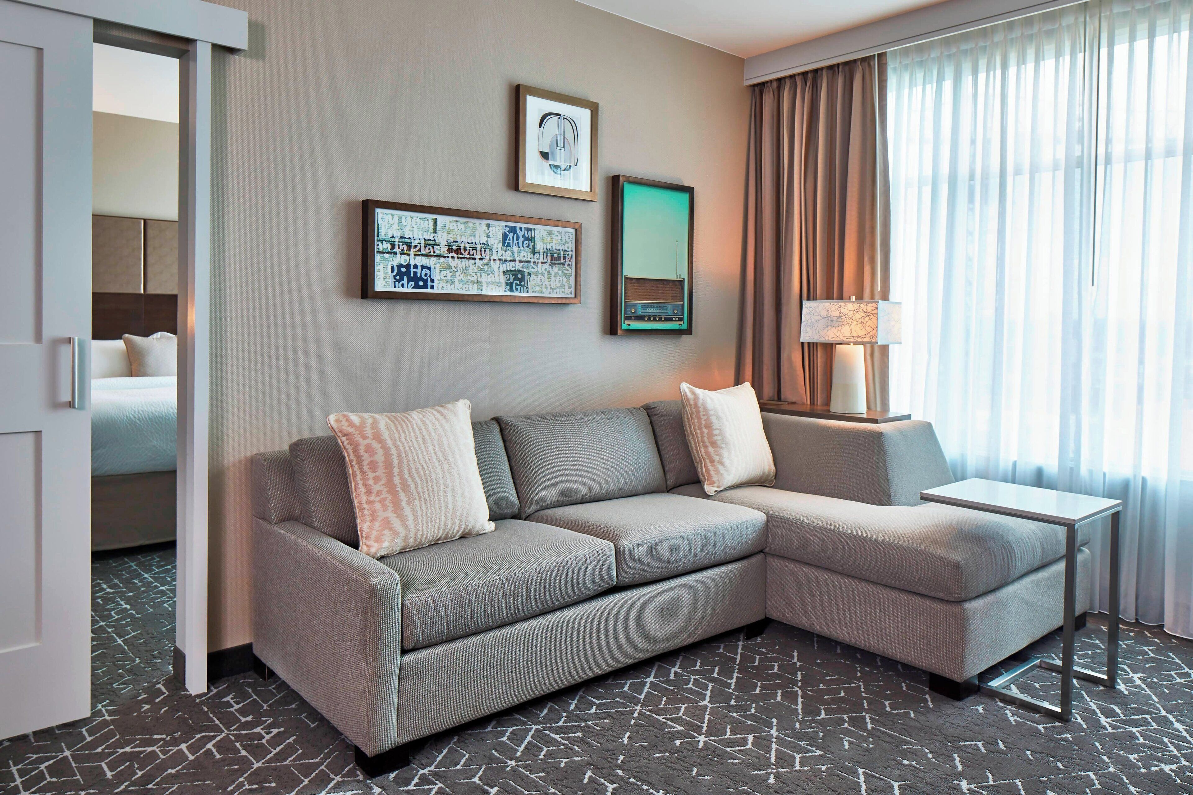Une chambre d’hôtel moderne avec un canapé d’angle gris, une petite table d’appoint, une œuvre d’art encadrée représentant un paysage urbain, une radio et une grande fenêtre avec des rideaux.