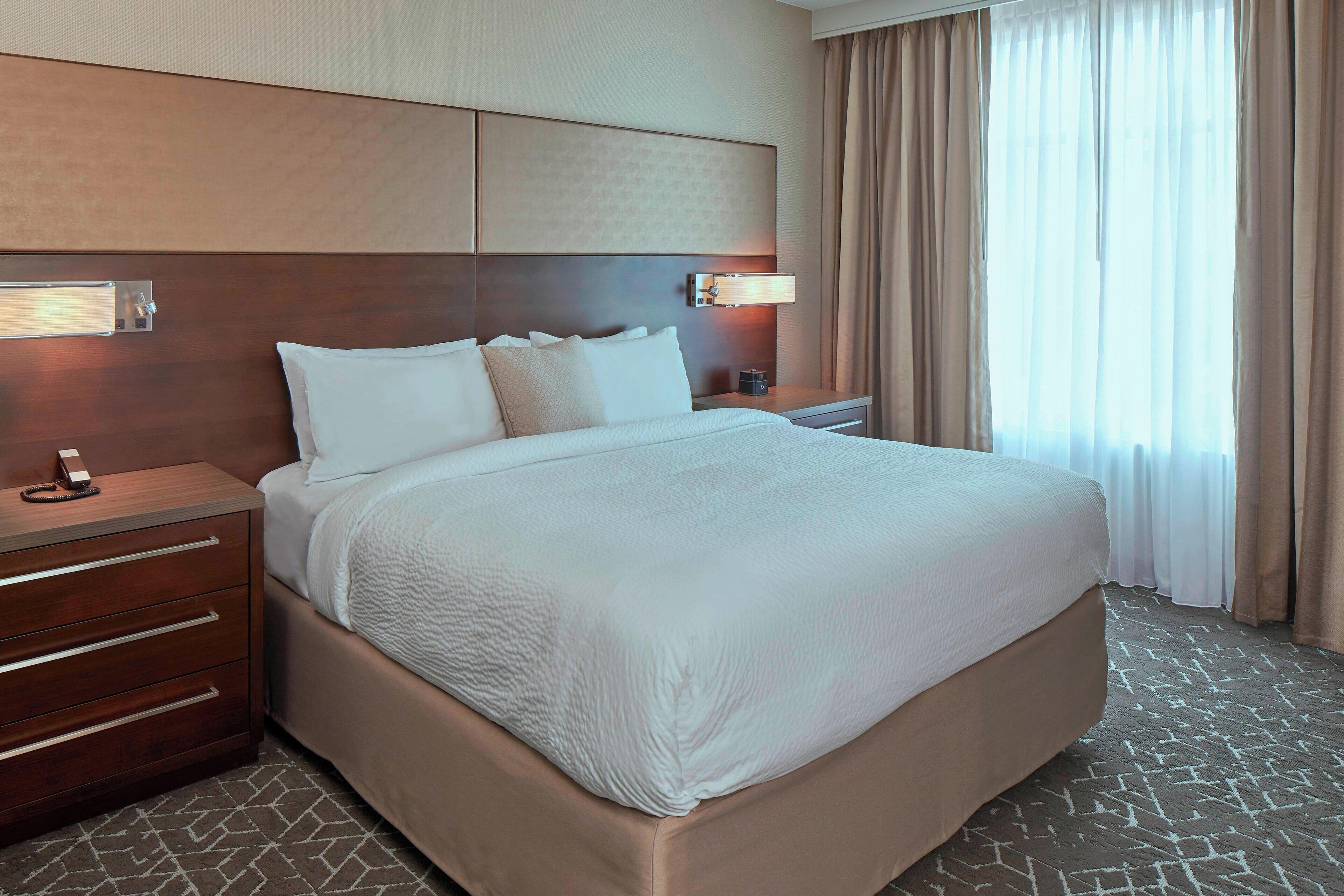 A hotel room with a large bed, a wooden headboard, a bedside table with a lamp, and a view of the outdoors through sheer curtains.