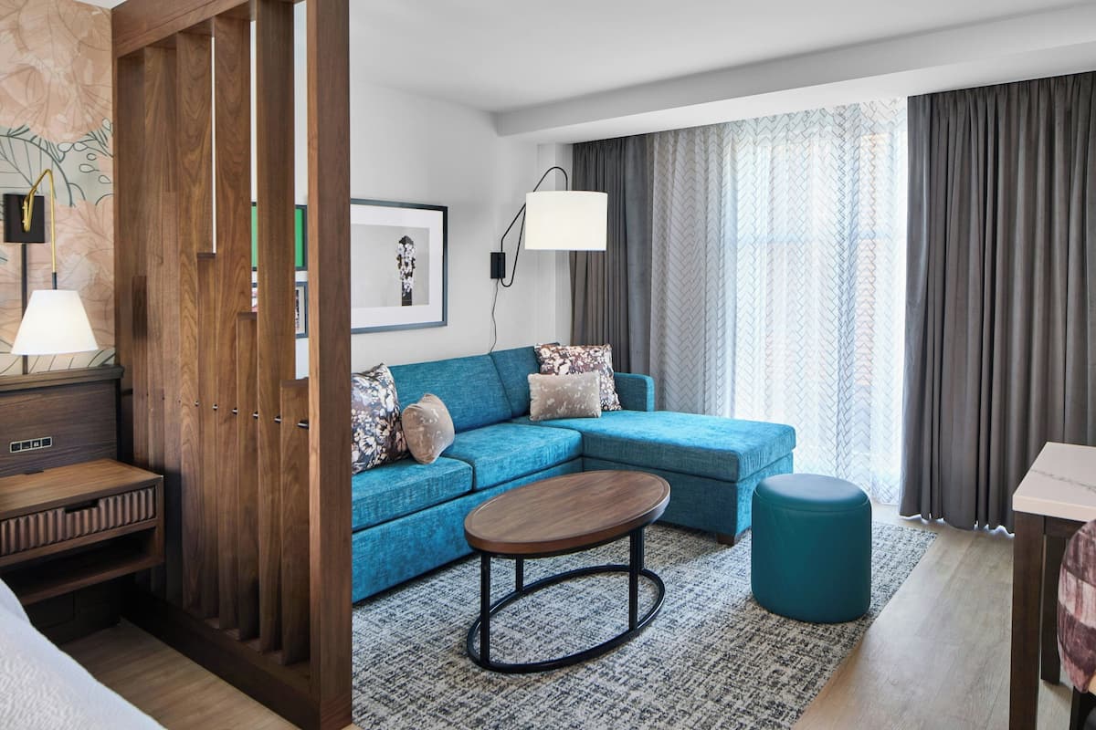 Una habitación de hotel moderna con sofá modular, una mesa redonda de centro, un armario de madera, un escritorio y un ventanal con cortinas.