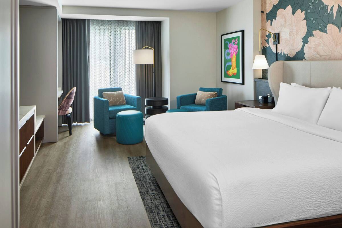 Habitación de hotel con una cama grande, dos sillones azules, una mesita redonda pequeña, un escritorio y un guardarropa.