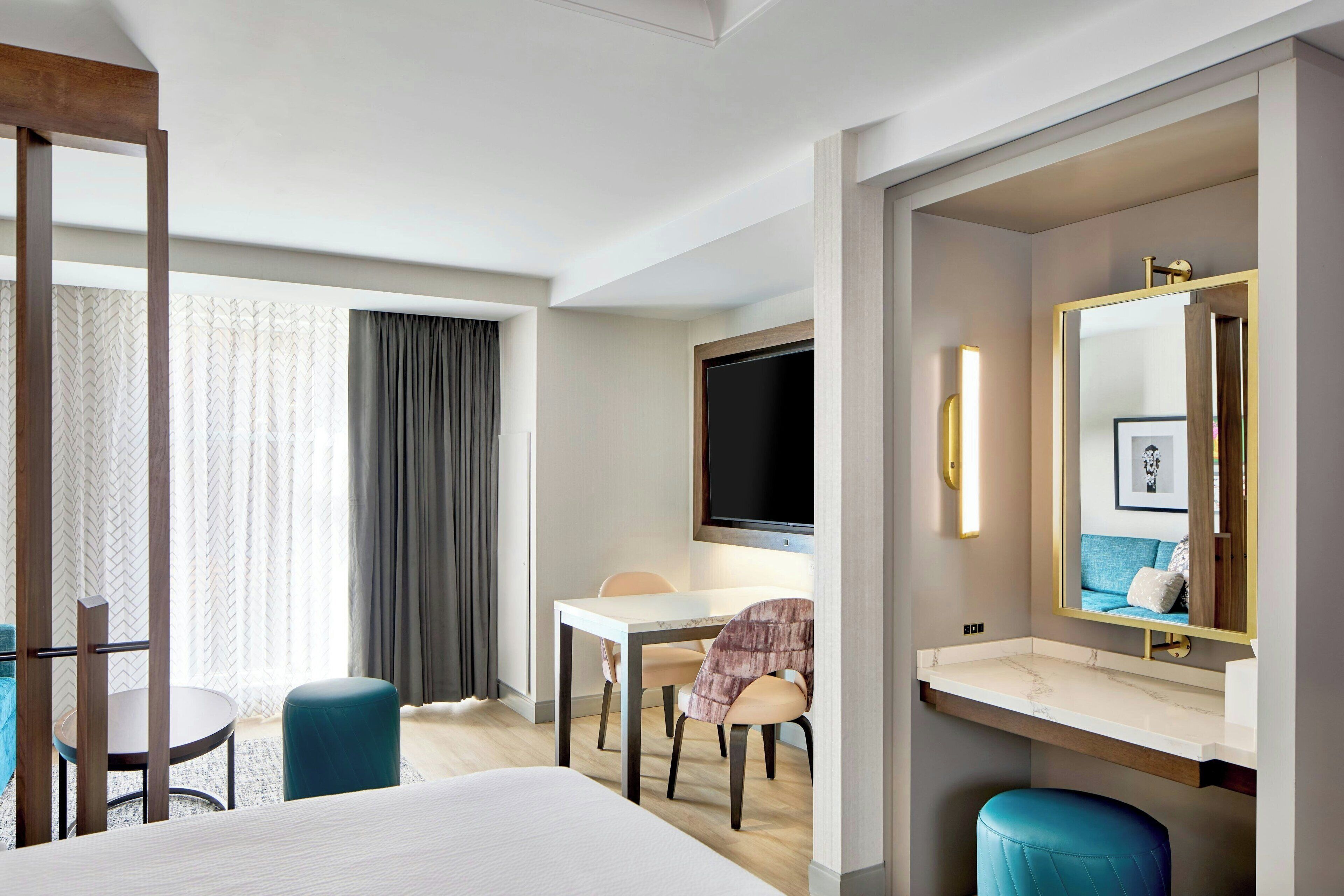 Quarto de hotel com cama, escrivaninha, cadeira, televisão e banheiro com espelho.