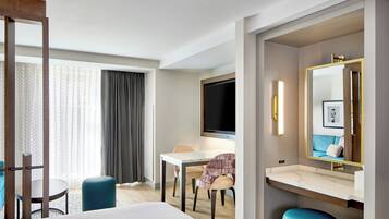 Quarto de hotel com cama, escrivaninha, cadeira, televisão e banheiro com espelho.