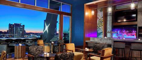 Zona de bar con asientos, vista a la ciudad y barra con botellas.