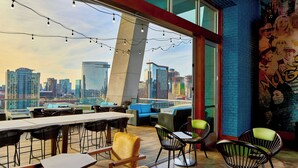 Un bar en la azotea con mesas y sillas, vista al horizonte de la ciudad y un mural en la pared.