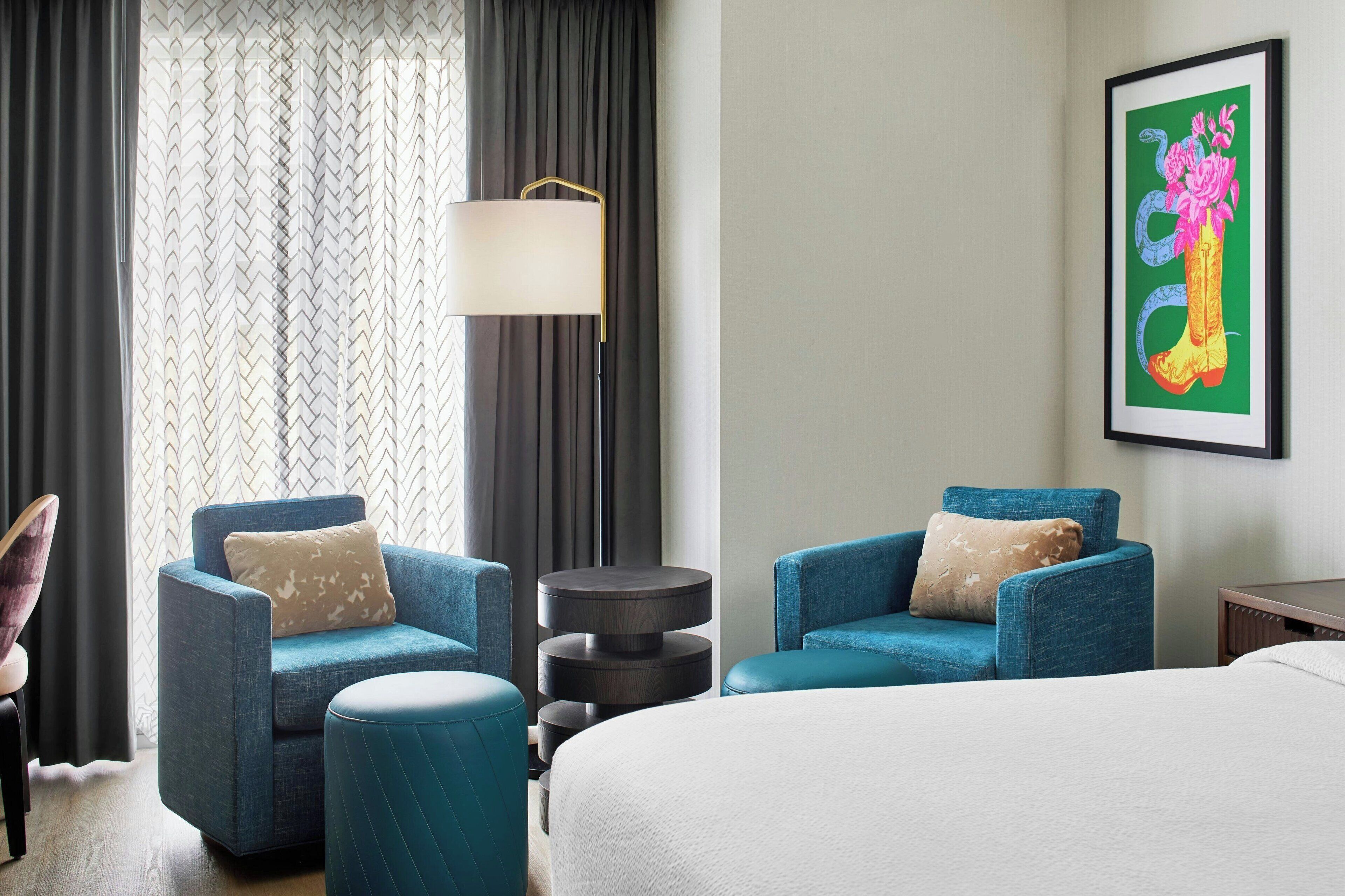 Habitación de hotel con dos sillones azules, una mesita redonda, una cama con ropa de cama blanca y una obra enmarcada en la pared.