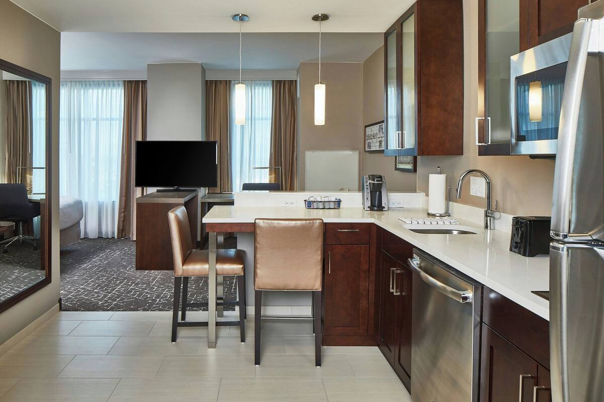 Habitación de hotel con kitchenette, comedor con mesa y sillas, televisor de pantalla plana, espejo grande y ventana con cortinas.