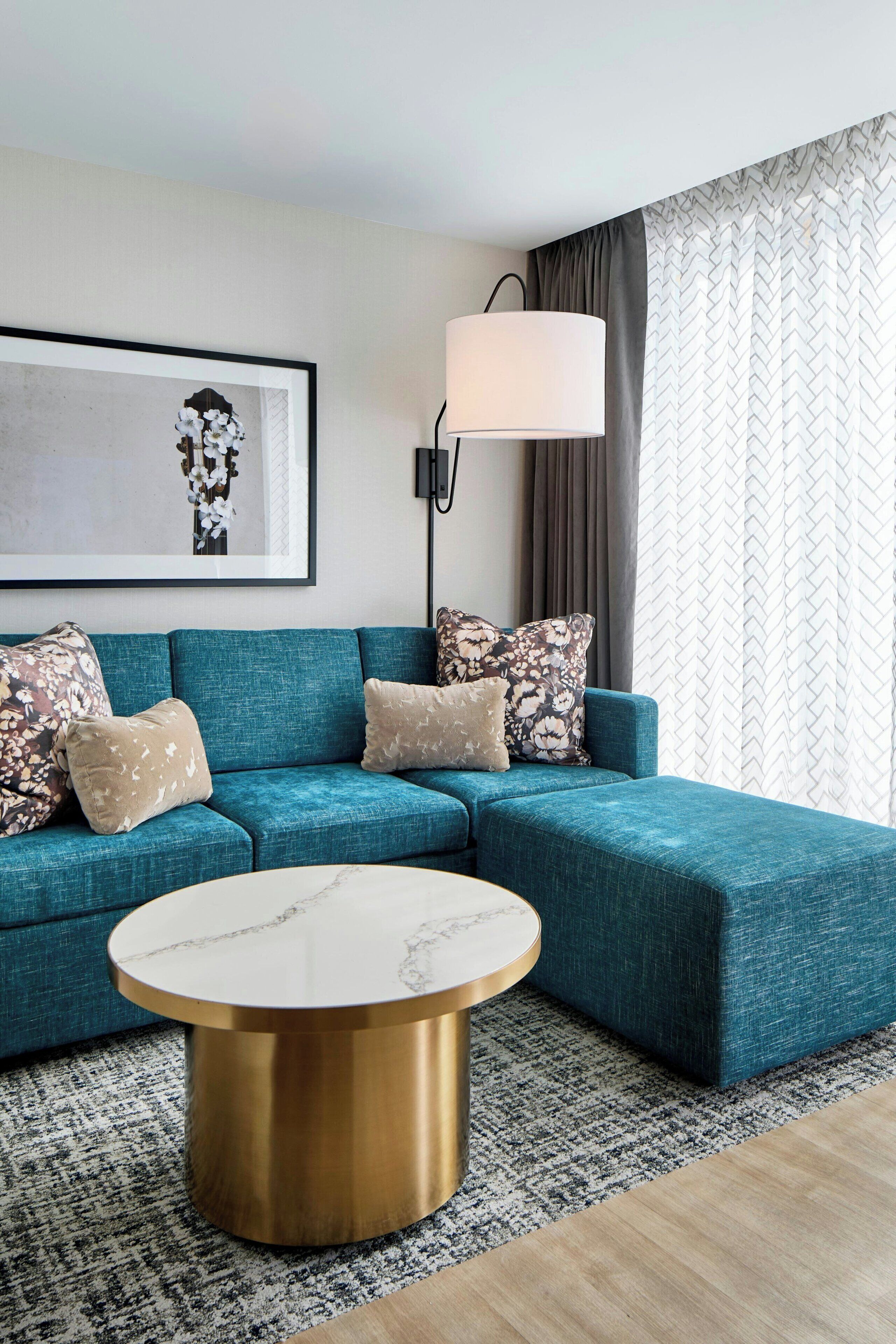 A modern living room with a teal sectional sofa, a round marble coffee table, a framed artwork, a floor lamp, and patterned curtains.