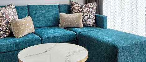 A modern living room with a teal sectional sofa, a round marble coffee table, a framed artwork, a floor lamp, and patterned curtains.