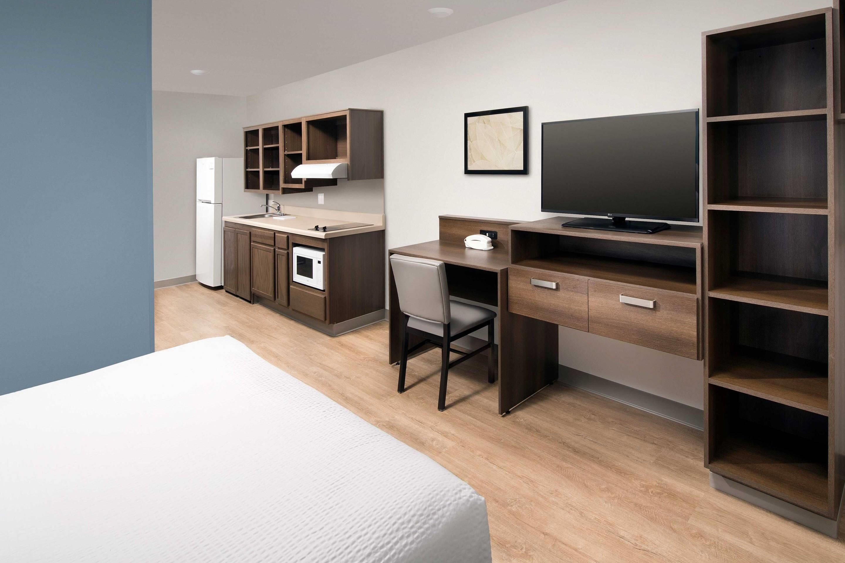 Une chambre d’hôtel moderne équipée d’une kitchenette, d’un lit, d’un bureau avec une chaise, d’une télévision et d’étagères intégrées.