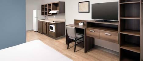 Une chambre d’hôtel moderne équipée d’une kitchenette, d’un lit, d’un bureau avec une chaise, d’une télévision et d’étagères intégrées.
