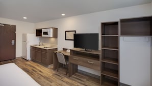 A compact hotel room with a kitchenette, a bed, a desk with a chair, a television, and built-in shelving.