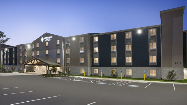 A modern hotel with a prominent sign reading "Woodspring Suites." It features a large parking lot with marked spaces and a well-lit entrance.