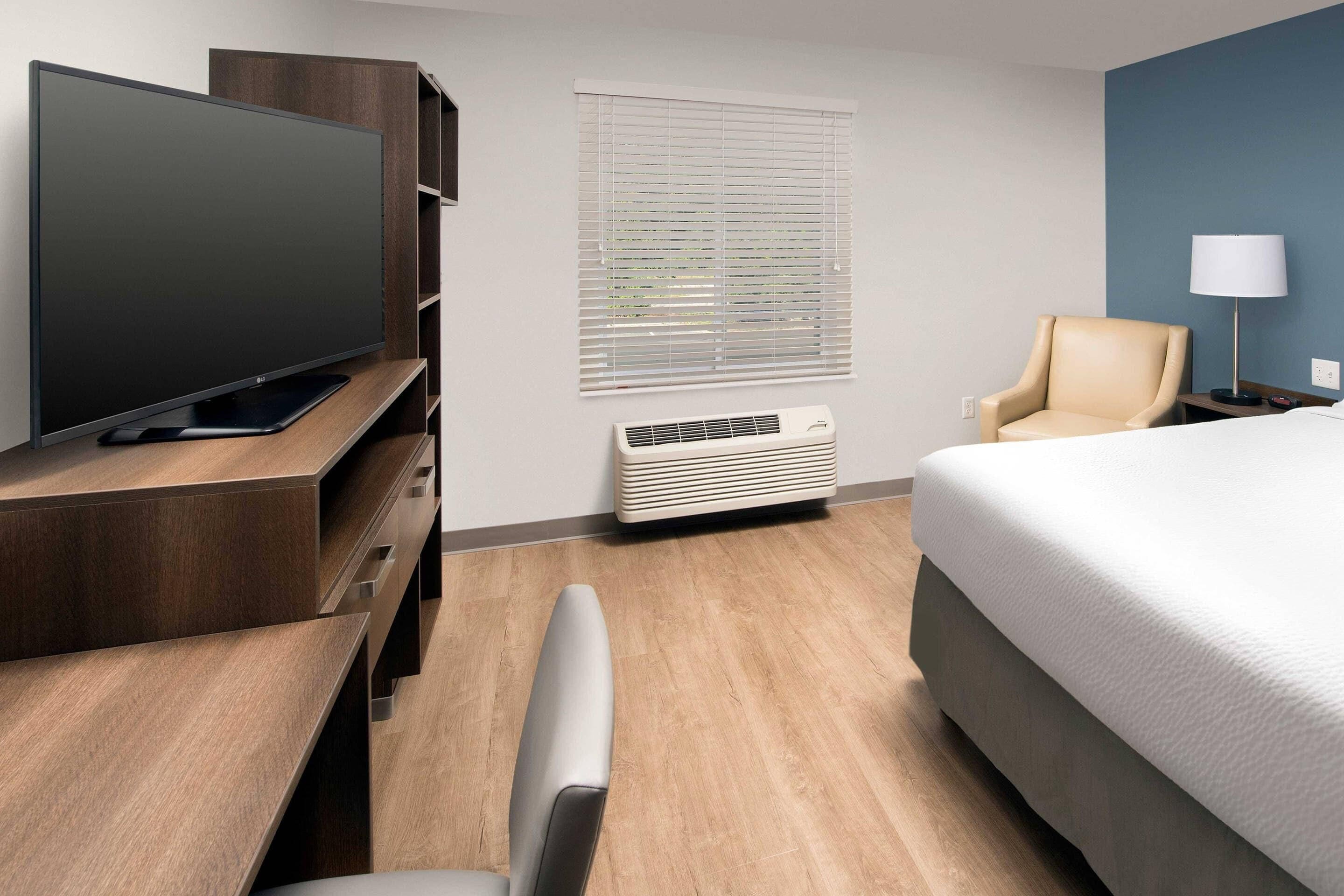 A hotel room with a flat-screen TV on a wooden stand, a bed with a white mattress, a chair, a desk with a computer, a window with blinds, and a wall-mounted air conditioner.