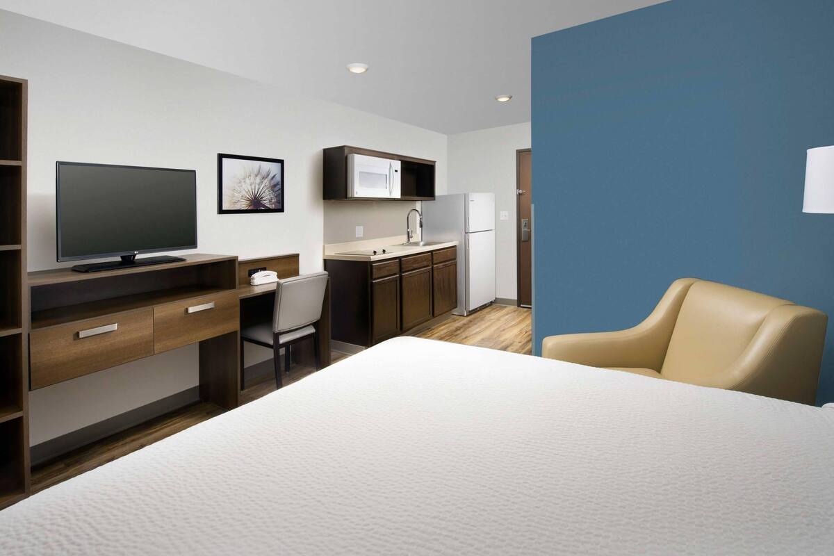 Ein modernes Hotelzimmer mit Bett, Schreibtisch, Sessel, Fernseher und einer Küchenzeile, sowie einer blauen Akzentwand.
