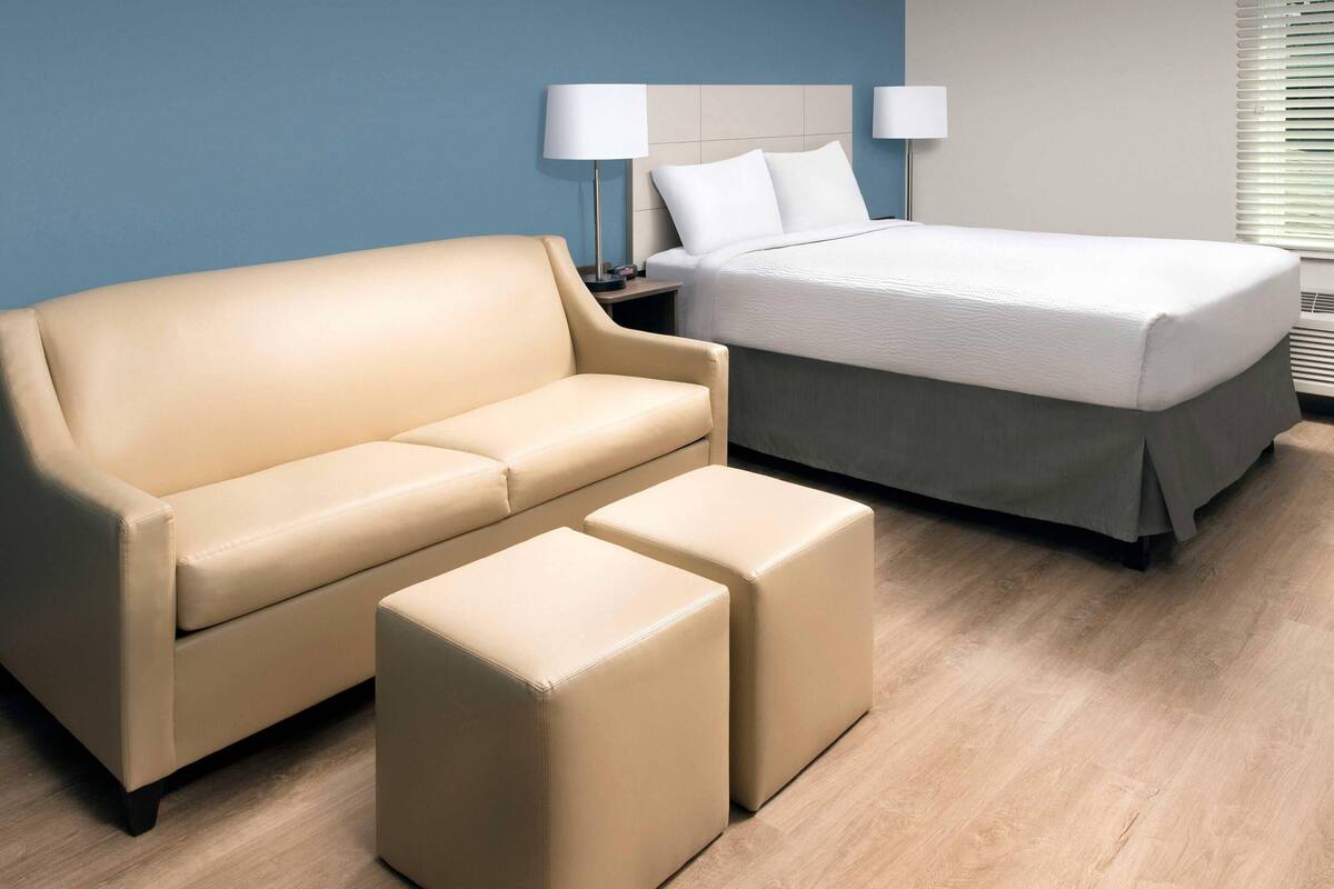 Ein Hotelzimmer mit beigefarbenem Sofa, zwei passende Poufs, einem Bett mit weißer Bettwäsche und einem Nachttisch mit Lampen.