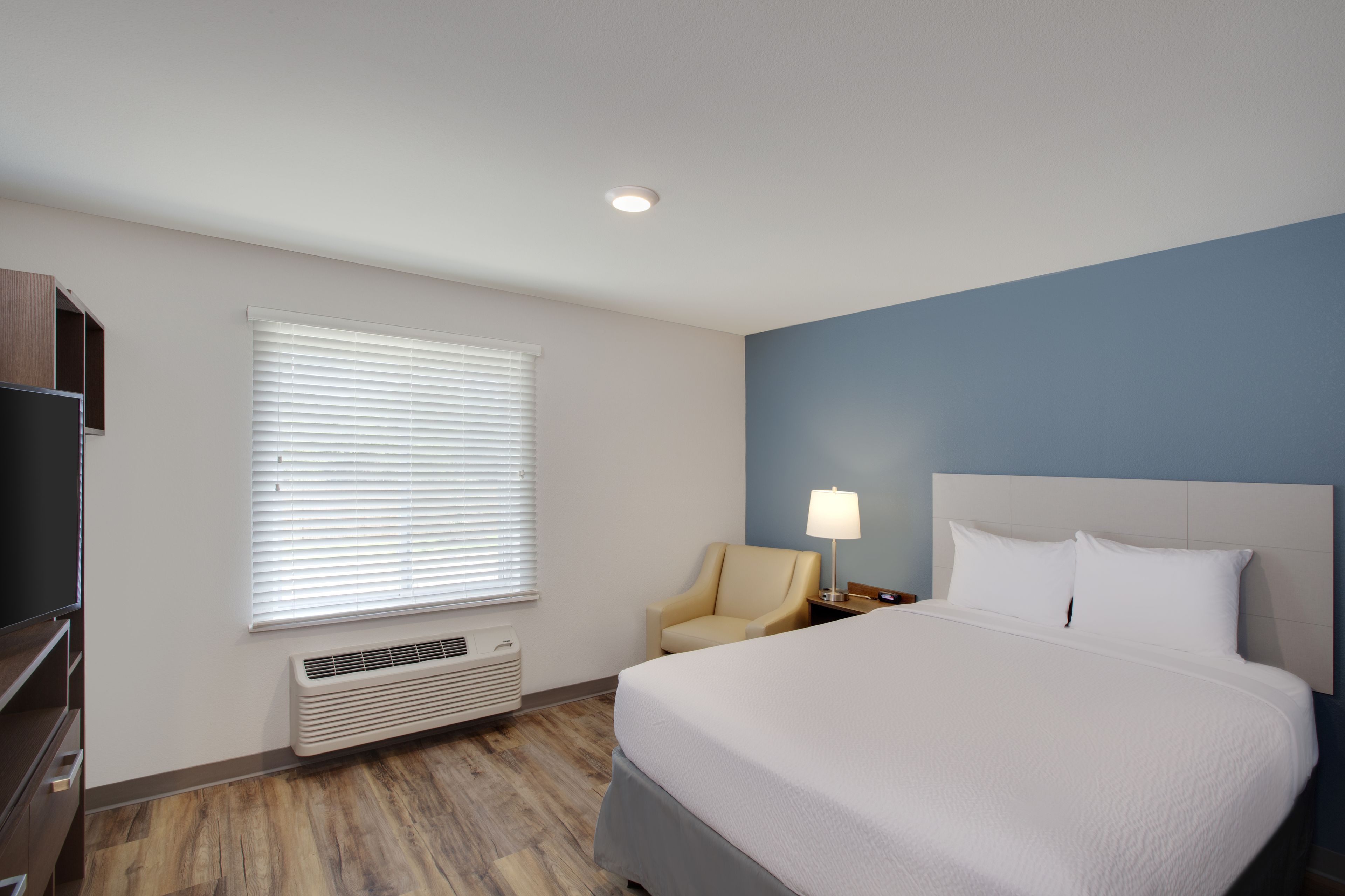 Une chambre d’hôtel avec un lit, une chaise, une télévision et une fenêtre avec des stores.
