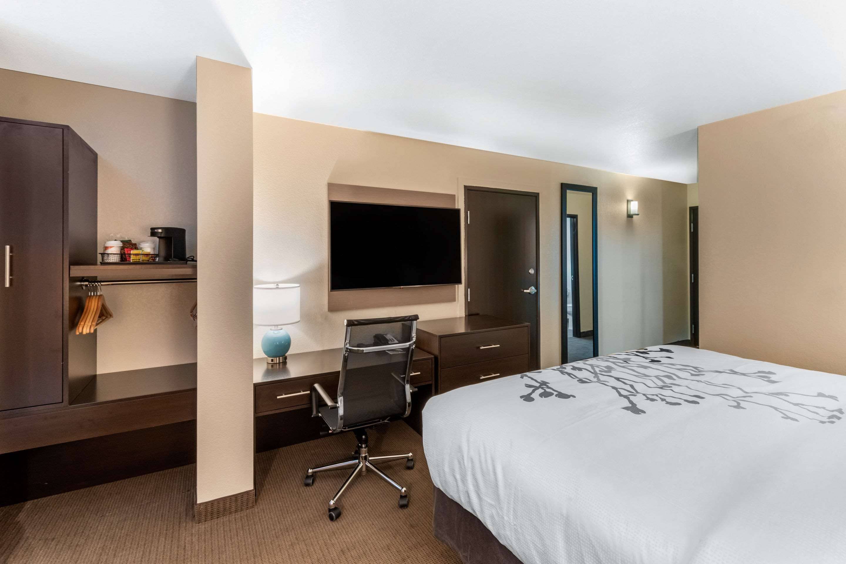 Habitación de hotel con cama, escritorio, silla, televisor, armario y pequeña área de kitchenette.