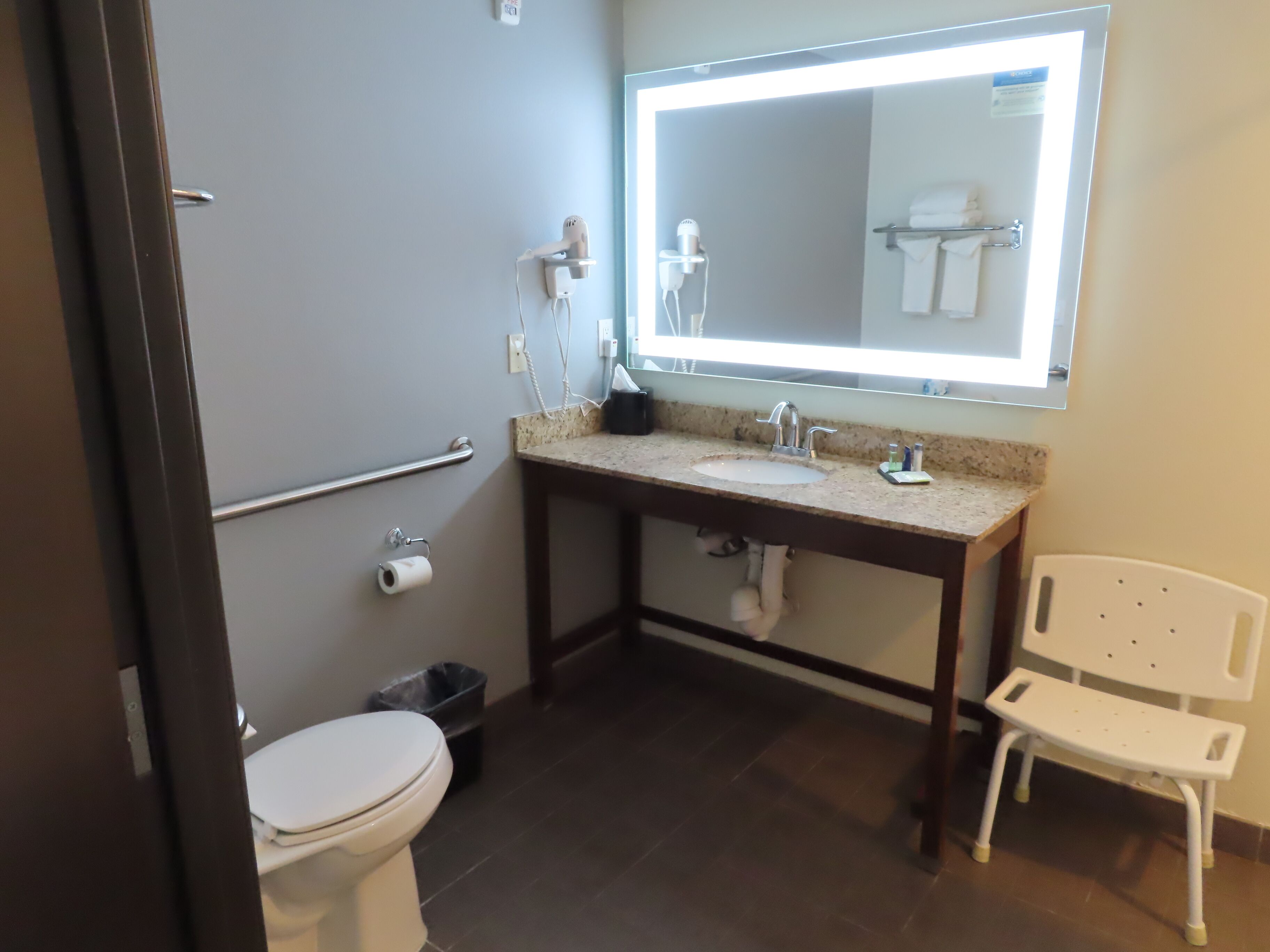 A bathroom with a toilet, sink, vanity, mirror, towel rack, and a white chair.