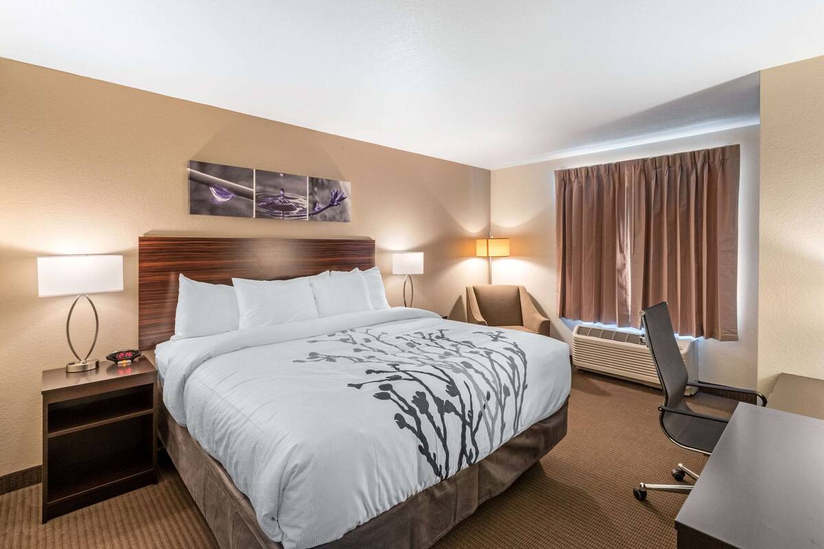 Ein Hotelzimmer mit einem großen Bett, zwei Nachttischen, einem Schreibtisch und einem Sessel. Zwei Wandleuchten und ein Fenster mit Vorhängen sind vorhanden.