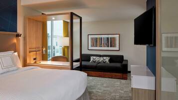 A hotel room with a large bed, a desk, a sofa, a TV, and a window with a view of buildings.