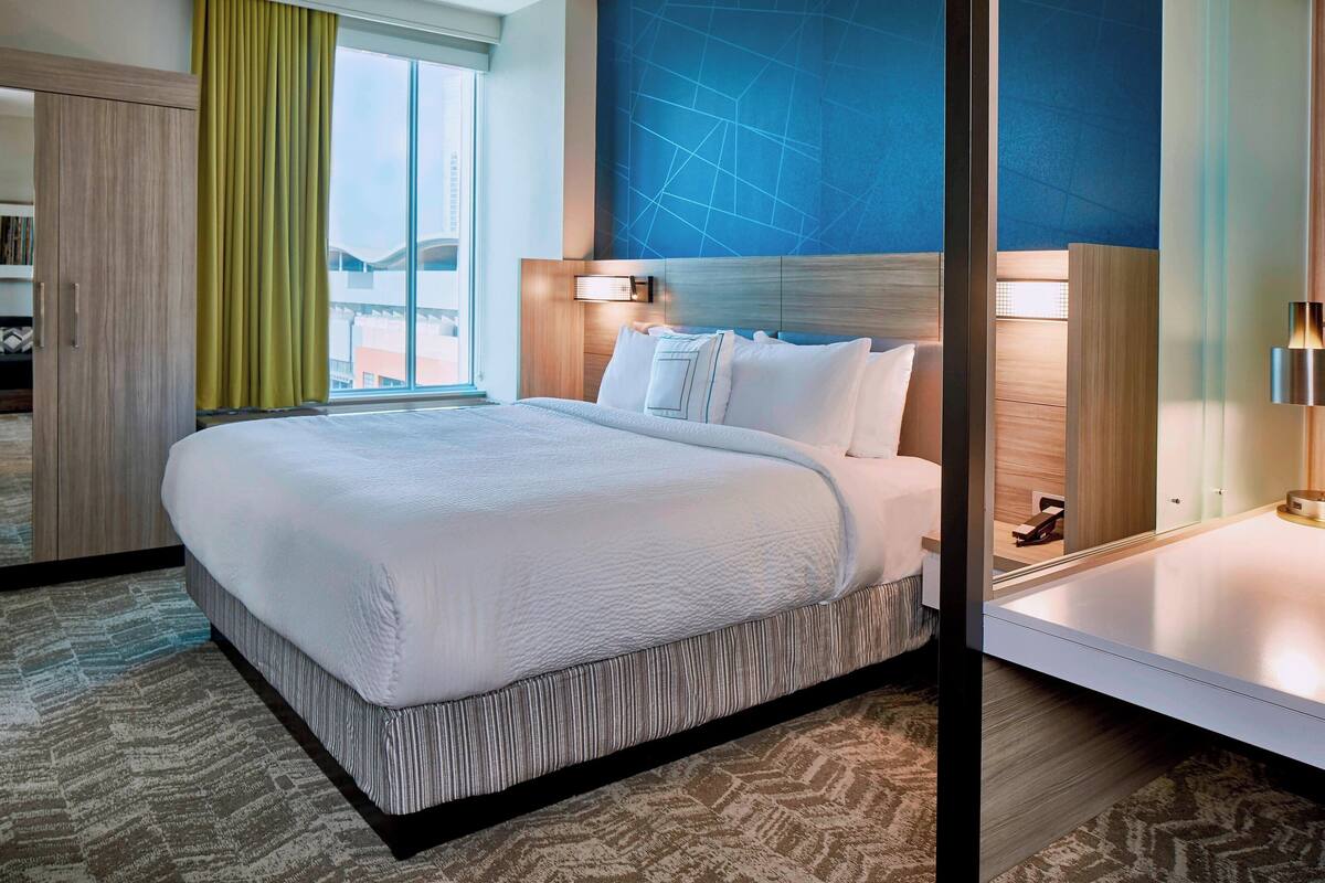 Une chambre d’hôtel avec un grand lit, une tête de lit en bois, une grande fenêtre avec des rideaux et un luminaire fixé au mur.