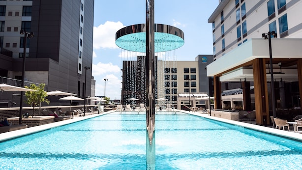 Una piscina con una fuente central, rodeada de sillas de descanso y sombrillas, con edificios altos al fondo.