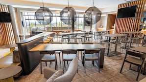 Un restaurante moderno con barra central, varias zonas de estar y grandes ventanales que dan a un aparcamiento.